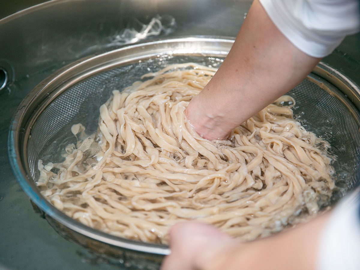 茹で上がったうどんを水に晒して滑りをとる。うどんは手打ちの平打ち縮れ麺。なのでつけ汁もよく絡む（食楽web）