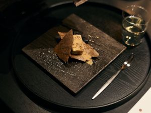 「聴く」テーマにした一皿。炭火の香りをまとったごま豆腐。皿の下に敷かれた玉砂利が擦れ合う音は、余市蒸溜所で守り続けられてきた石炭直火蒸溜の音と重なります[食楽web]